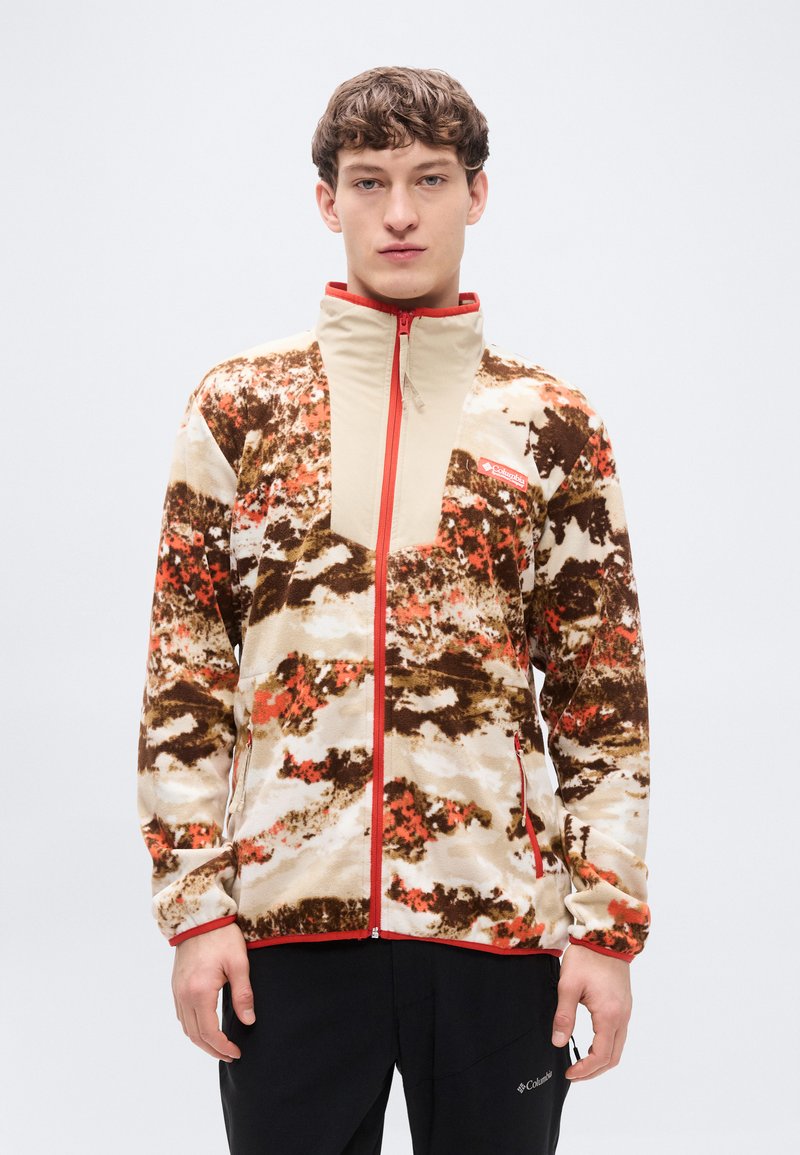 Ζακέτα από φλις με καμουφλάζ σχέδιο σε μπεζ, καφέ και πορτοκαλί, με αντίθετη μονή μπροστινή επένδυση σε ταμπά και κόκκινες λεπτομέρειες φερμουάρ.