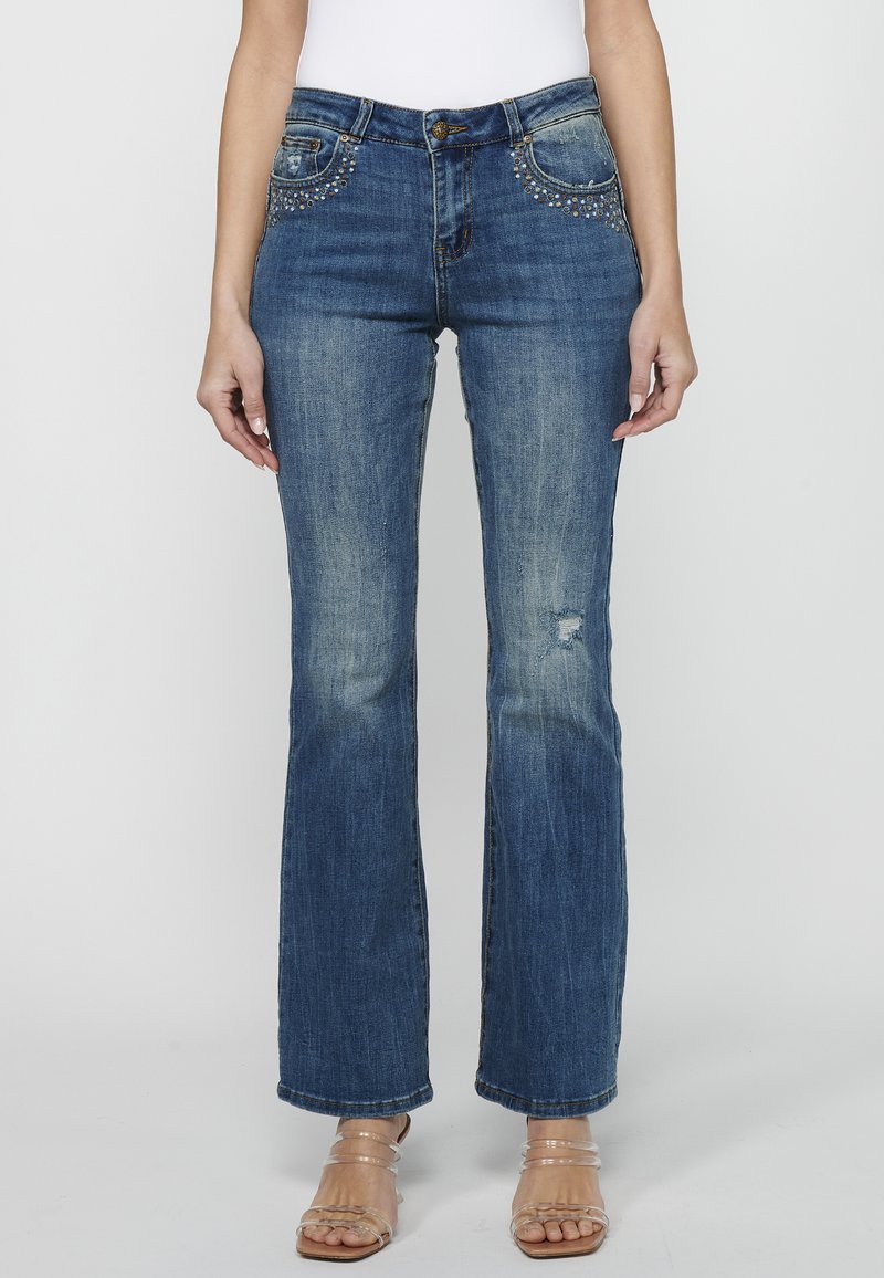 Jeans de mezclilla azul de pierna acampanada con una cintura de altura media, detalles desgastados y bolsillos decorados con tachuelas. El material tiene una ligera textura.