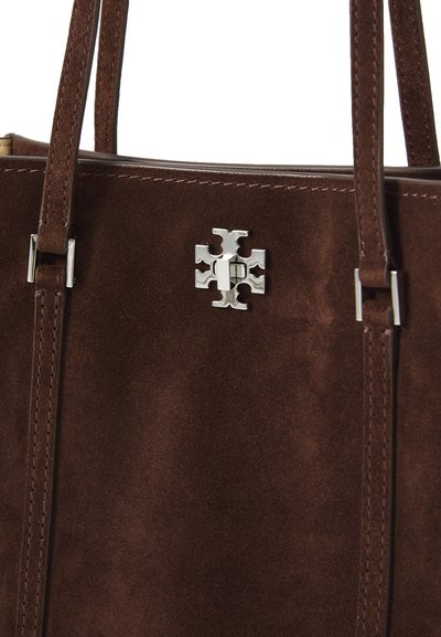 Brun ruskinds tote bag med dobbelte håndtag, prydet med et sølvfarget korslogo og kontrastsyning. Glat tekstur og robust design.