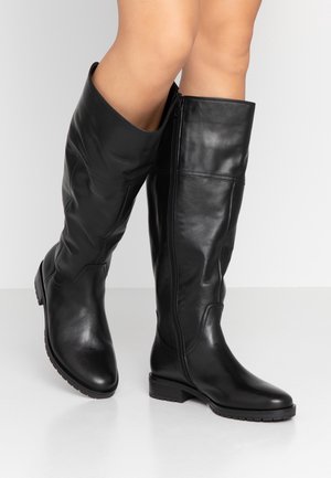 Widok nóg osoby noszącej czarne skórzane kozaki do kolan z niskimi obcasami i zamkami błyskawicznymi po bokach na białym tle.