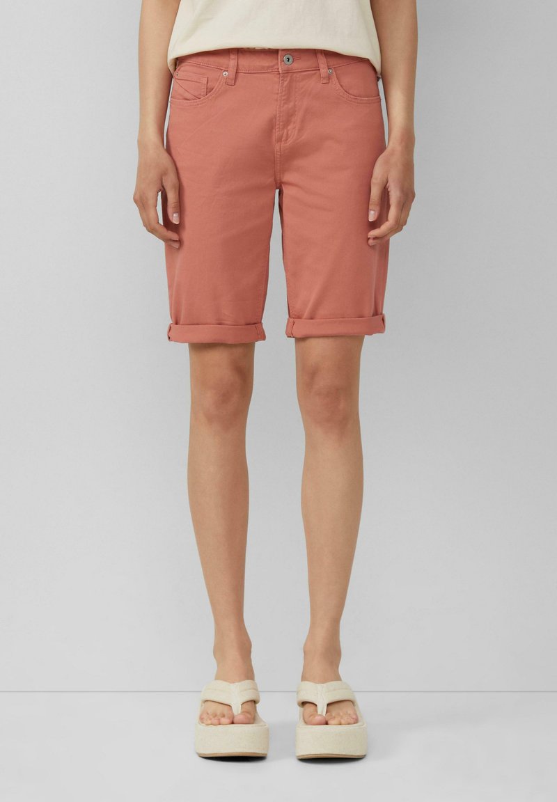Shorts en denim rose avec ourlets retroussés, présentant un style à cinq poches et une fermeture à bouton en métal. Portés avec des sandales à plateforme blanc cassé.