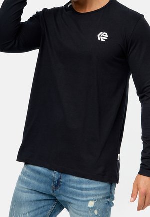 Uomo che indossa una camicia nera a maniche lunghe con un piccolo logo bianco sul petto e jeans azzurri strappati.