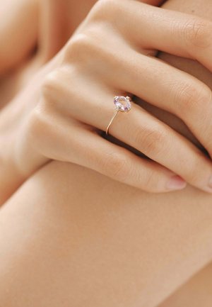 Anillo de oro con una gema ovalada de color lavanda, flanqueada por pequeñas piedras claras, sobre un fondo de tono de piel natural y suave.