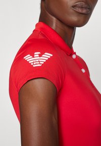 Rode polo shirt met een gektextureerde stof, korte mouwen en een wit arendlogo op de linkerschouder. Drie knopen bij de kraag.