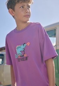 Paarse katoenen T-shirt met een chameleonafbeelding op een scooter en de tekst "KEEP ROLLIN'". Korte mouwen en een ronde hals.