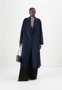 Cămașă lungă, bleumarin, confecționată dintr-un amestec de lână. Design cu două rânduri de nasturi mari, negri. Asortată cu pantaloni largi negri și un top texturat bej.
