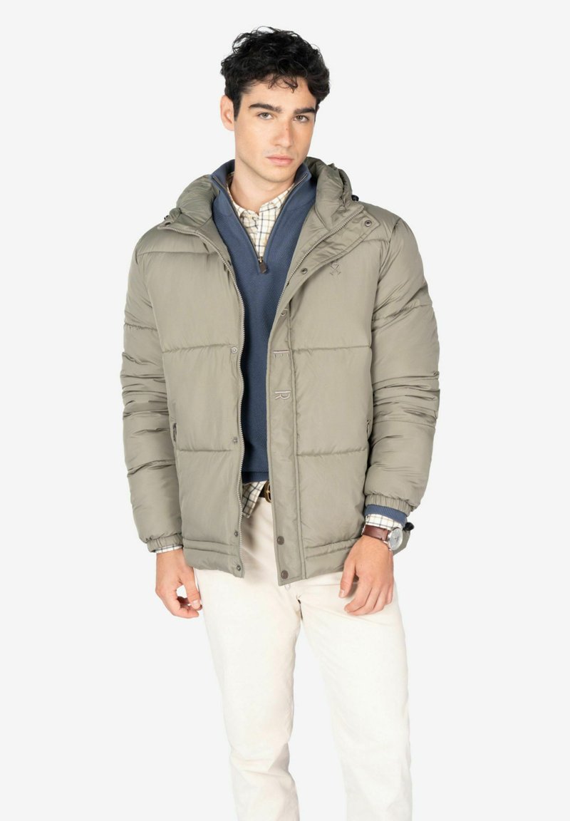 Giacca puffer oliva chiara con zip frontale, cappuccio attaccato e polsini elasticizzati, indossata sopra un maglione blu navy e una camicia a quadri.