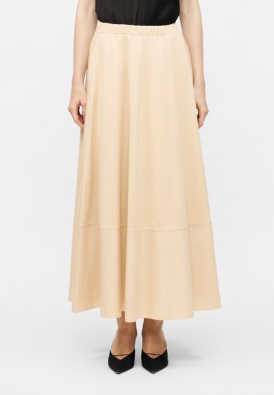 Femme portant une jupe beige longueur cheville avec une taille élastique et des escarpins noirs à bout pointu, debout devant un fond blanc uni.
