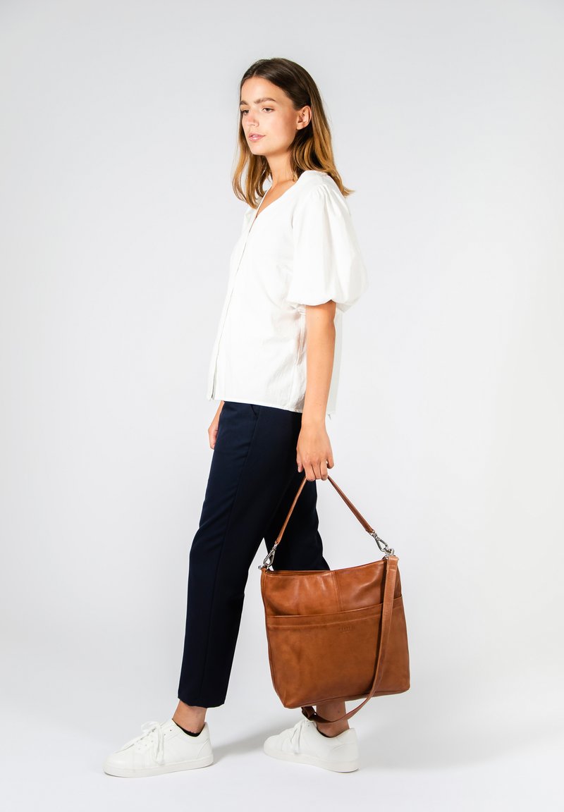 Borsa a spalla in pelle marrone chiaro con texture liscia, accessori in argento e una singola tracolla regolabile, tenuta da una persona con una maglietta bianca e pantaloni blu navy.