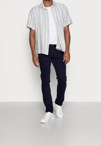 Camisa de manga corta a rayas en azul y blanco, combinada con vaqueros oscuros y zapatillas blancas. La camisa presenta un cuello clásico y botones.