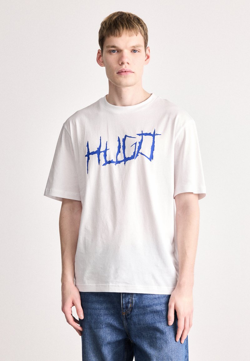 Un tricou alb din bumbac cu un text grafic mare, albastru, "HUGO" într-o font jagged. Mâneci scurte și croială lejeră, asortat cu blugi albaștri.