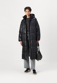 Abrigo acolchado largo negro con capucha, diseño acolchado, textura brillante y cremallera de longitud completa. Llevado con jeans y zapatillas.