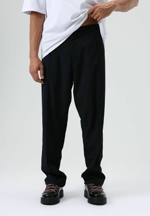 Zwarte geplooide broek met een ontspannen pasvorm, gemaakt van lichtgewicht stof. Gecombineerd met zwarte schoenen met patroonveter.