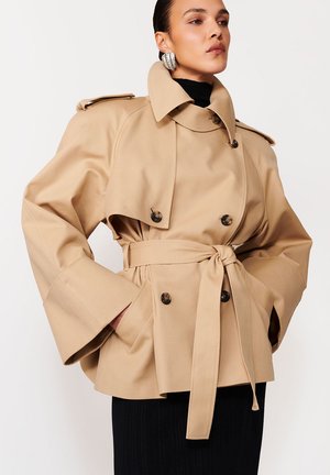Kobieta w beżowym płaszczu trencz z paskiem, szerokimi rękawami i dużymi guzikami, w połączeniu z czarną prążkowaną spódnicą i efektownymi kolczykami.