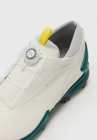 Vit sportskor med en texturerad ovandel, BOA-snörningssystem, gul detalj vid hälen och mörk teal-accenter på sulan.