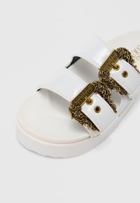 White patent slide sandal featuring two gold-tone decorative buckles, textured footbed, and a thick white sole.