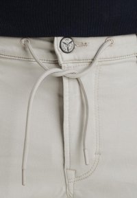 Beige pants with a drawstring closure, metal button featuring a logo, and contrasting stitching; smooth texture and fitted design.