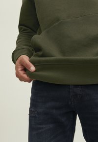 Olivgrüner Sweatshirt mit geripptem Saum und Bündchen, kombiniert mit dunkler Jeans, die beschädigte Stellen aufweist. Eine Hand hält den Saum des Sweatshirts.