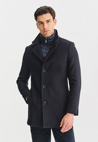 Czarny, wełniany płaszcz w kratę z klapami, czterema guzikami i kontrastującą niebieską kamizelką puchową pod spodem. Gładka tekstura.