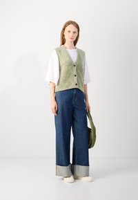 Groene, fuzzy gebreide vest met vijf knopen, gelaagd over een wit topje. Gepaard met een wide-leg, donkerblauwe jeans met lichtgrijze boorden.