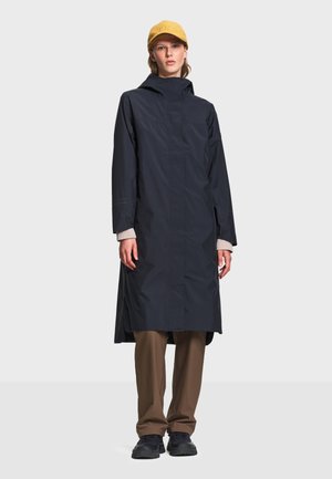 Personne portant un long manteau bleu marine, un pantalon marron, des chaussures noires et une casquette jaune, debout devant un fond gris clair.