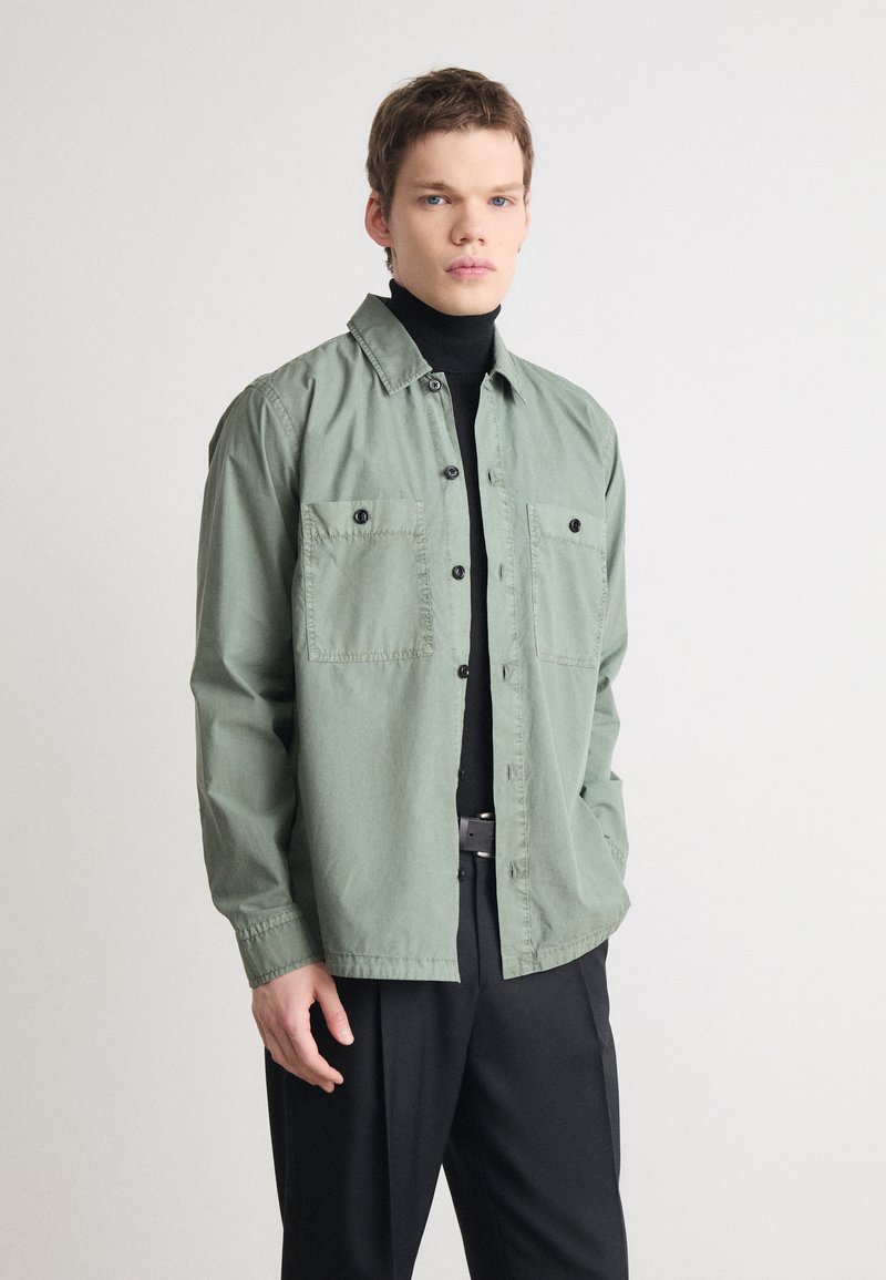 Camisa de botones verde claro con dos bolsillos en el pecho, de tela ligera, de manga larga y con un corte relajado, llevada sobre un cuello de tortuga negro.