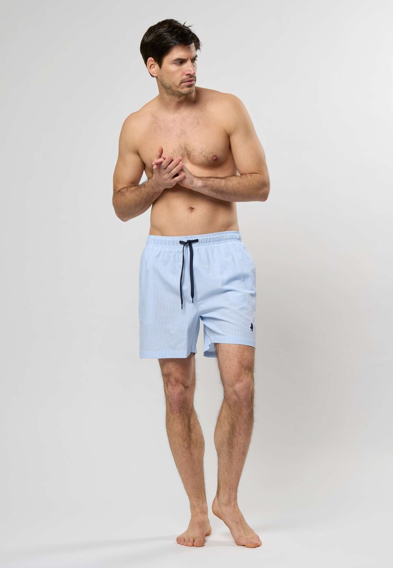 Homme torse nu, debout pieds nus, portant un short de bain bleu clair avec un cordon noir, regardant vers sa gauche sur un fond uni.