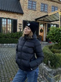 Svart dunjakke med horisontale polstringer, glidelåslukking og sidelommer. Brukt med en svart strikket lue. Bakgrunn med steinbelagt underlag.