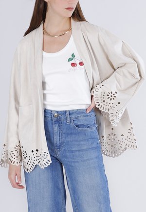 Mujer con chaqueta beige con bordes florales cortados con láser sobre una camiseta sin mangas blanca bordada con flores de cerezo y vaqueros azules, con la mano apoyada en el bolsillo.