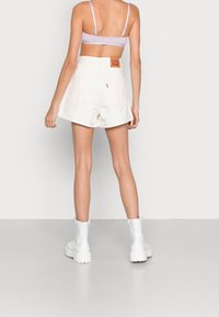 Mujer vistiendo pantalones cortos de tiro alto Levi's blancos, bralette lila y botas blancas hasta el tobillo, de pie contra un fondo gris claro liso.