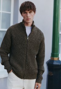 Ritsvest in donkerolijfgroen, geribbelde textuur, hoge kraag en zijzakken; gedragen over een wit T-shirt en gecombineerd met lichte broek.