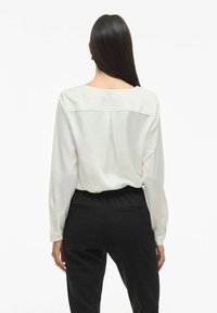 Blusa bianca a maniche lunghe con scollo sulla schiena e dettagli plissettati, abbinata a pantaloni neri con vita elastica e tasche posteriori.