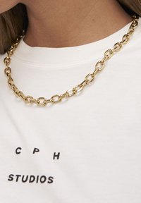 Guldhalskæde med store ovale led, båret over en hvid skjorte med sort trykt tekst. Glat, skinnende finish med et struktureret design.