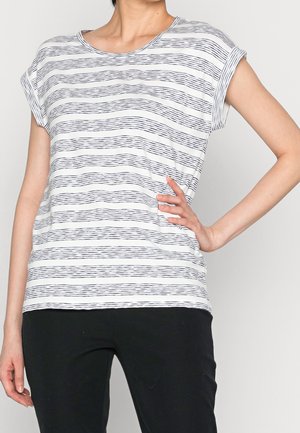 Femme portant un haut blanc à manches courtes avec de fines rayures horizontales noires et un pantalon noir, la main posée sur la hanche.