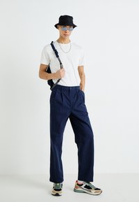 Navy pleated trousers with a high waist, paired with a white t-shirt, black bucket hat, and sunglasses. Worn with multi-colored sneakers.