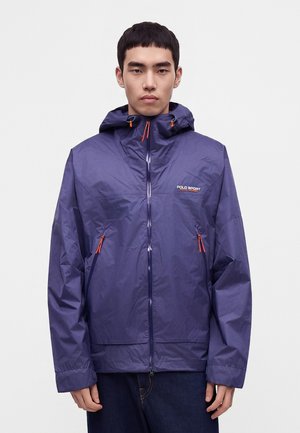 Joven con una chaqueta azul con capucha Polo Sport y tiradores de cremallera naranja, de pie contra un fondo claro y liso.