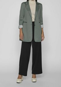 Zelené sako s klopovým límcem, černé kalhoty a světle béžový rolák. Má pruhované manžety na rukávech a robustní bílé platformové boty.