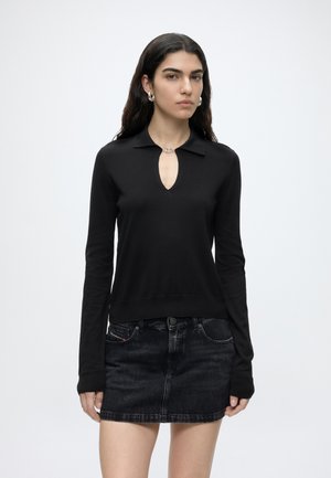Giovane donna con capelli lunghi e scuri che indossa un top nero a maniche lunghe con scollo a goccia e una minigonna di jeans scuro.