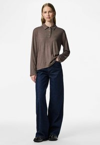 Žena stoji s rukom u džepu, odjevena u smeđu dugouhanu polo majicu, široke tamnoplave traperice i crne cipele na bijeloj pozadini.