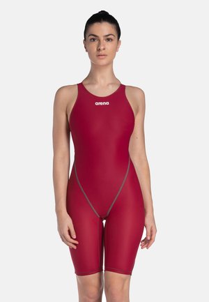 Nainen, joka käyttää hihattomaa, polvipituista punaista Arena-merkkistä uimapukua, jossa on harmaat saumat, seisoo yhtenäistä vaaleaa taustaa vasten.