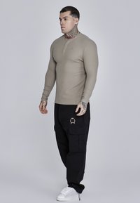 Bež majica dugih rukava s dugmadi, u kombinaciji s crnim cargo hlačama s zlatnim emblemom i bijelim tenisicama. Glatka tekstura tkanine.