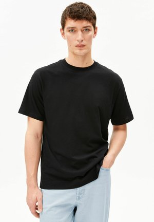 Jonge man die een eenvoudige zwarte T-shirt met korte mouwen en lichtblauwe jeans draagt, staand met zijn rechterhand in zijn broekzak.