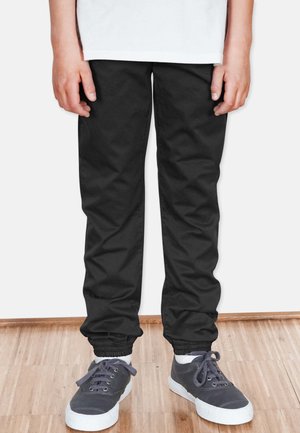 Schwarze Hose mit einer schmal zulaufenden Passform und elastischen Bündchen, aus einem glatten Stoff gefertigt. Getragen mit grauen Sneakers mit weißen Sohlen.