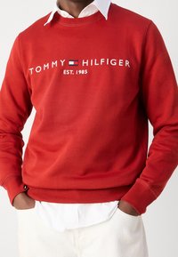 Czerwony sweter z bawełny, z okrągłym dekoltem, długimi rękawami i białym napisem "TOMMY HILFIGER EST. 1985".