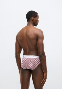Czerwone bokserki w geometryczny wzór z białym paskiem w talii z logo "Calvin Klein". Gładka tkanina z geometrycznym motywem. Model prezentowany od tyłu.