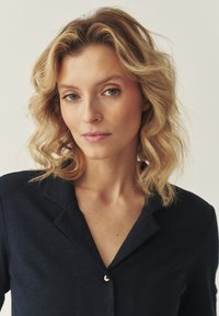 Cămașă bleumarin cu nasturi și guler, având o textură netedă și o strălucire subtilă. Păr blond, moale și ondulat, de lungime până la umeri.