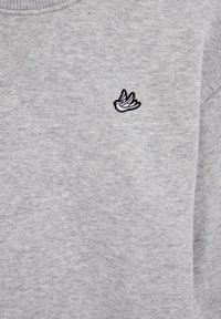 Szürke pulóver puha anyagból, bal mellrészen kis fekete hímzett madár logóval. A textúra sima és enyhén bolyhos hatású.