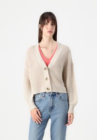 Beige strikket cardigan med dyp V-hals, tre store knapper og ballongsleeves, kombinert med en rosa topp og lyseblå jeans.