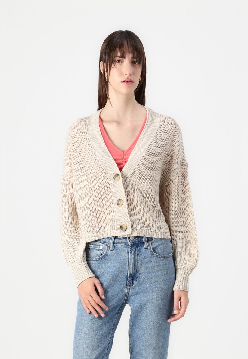 Beige strikket cardigan med dyp V-hals, tre store knapper og ballongsleeves, kombinert med en rosa topp og lyseblå jeans.