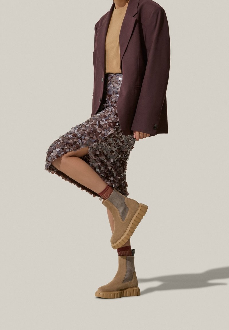 Personne portant un blazer marron oversize, un top beige, une jupe à sequins texturée, des chaussettes bordeaux et des bottines à plateforme beige, levant une jambe en plein pas.